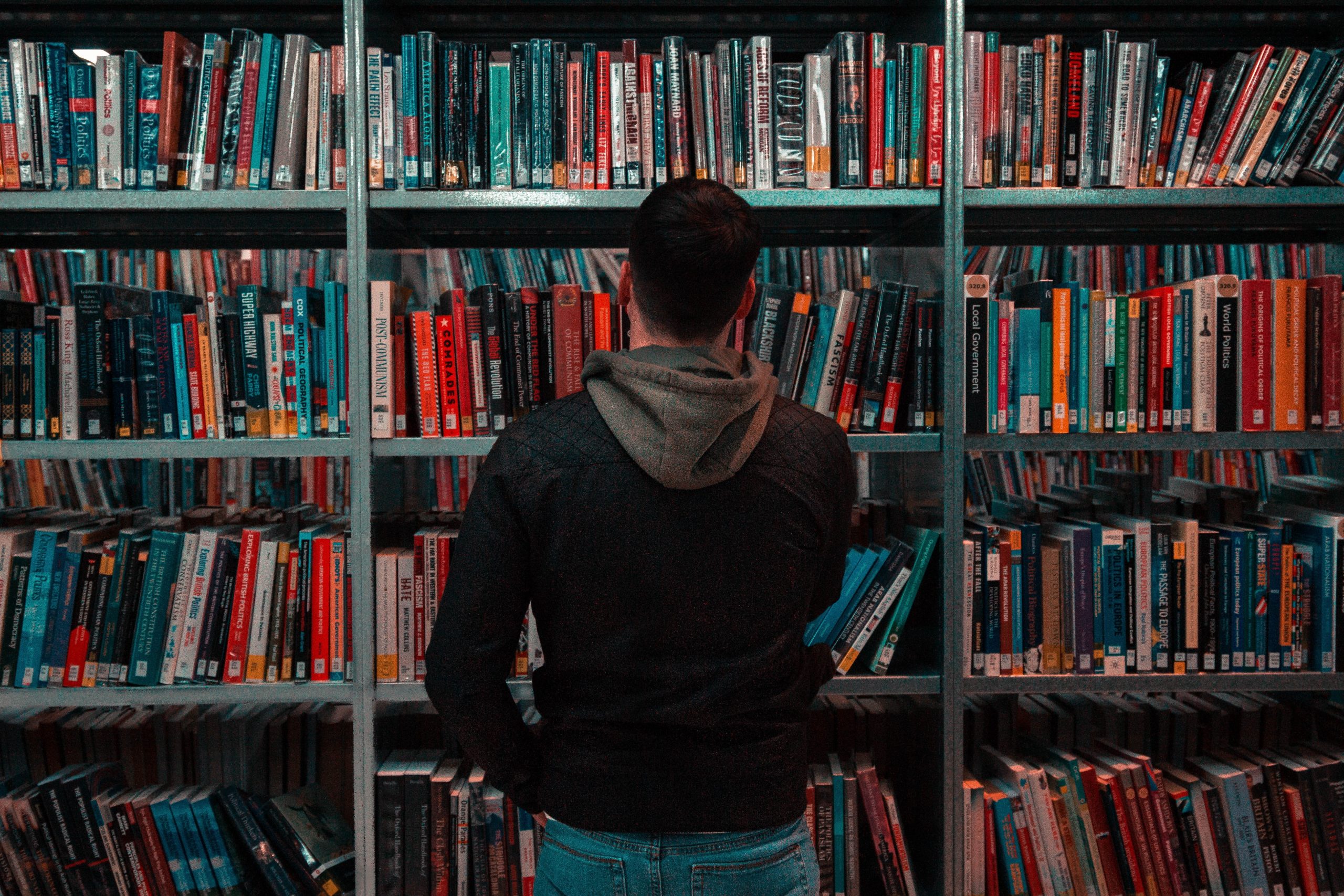 Image of a dark-lit bookshelf with a figure standing facing it. The figure wears a dark hoodie.
